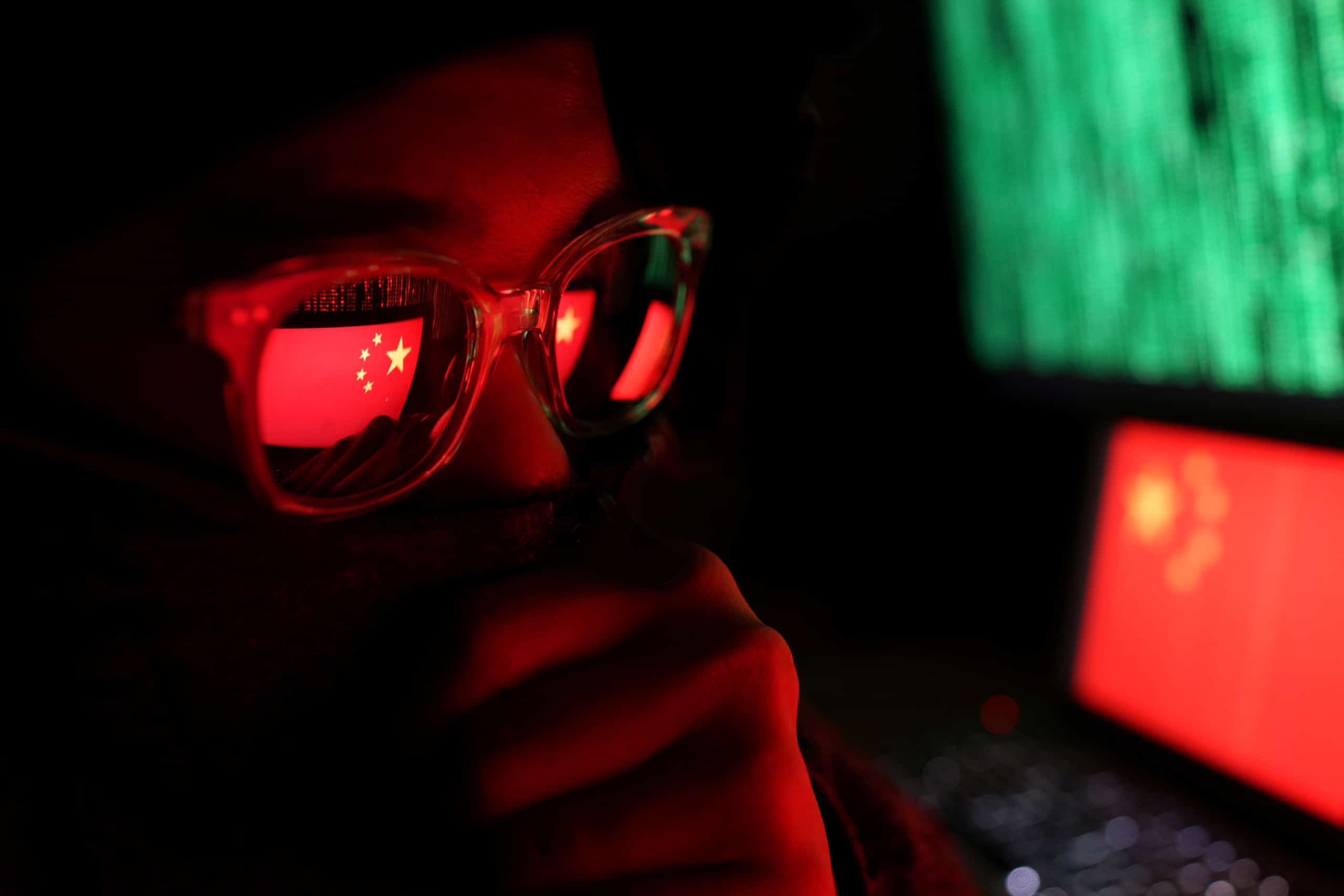 Person in a dark room wearing glasses with hand over their mouth to obscure face; reflected on their glasses is the China flag displayed on a laptop